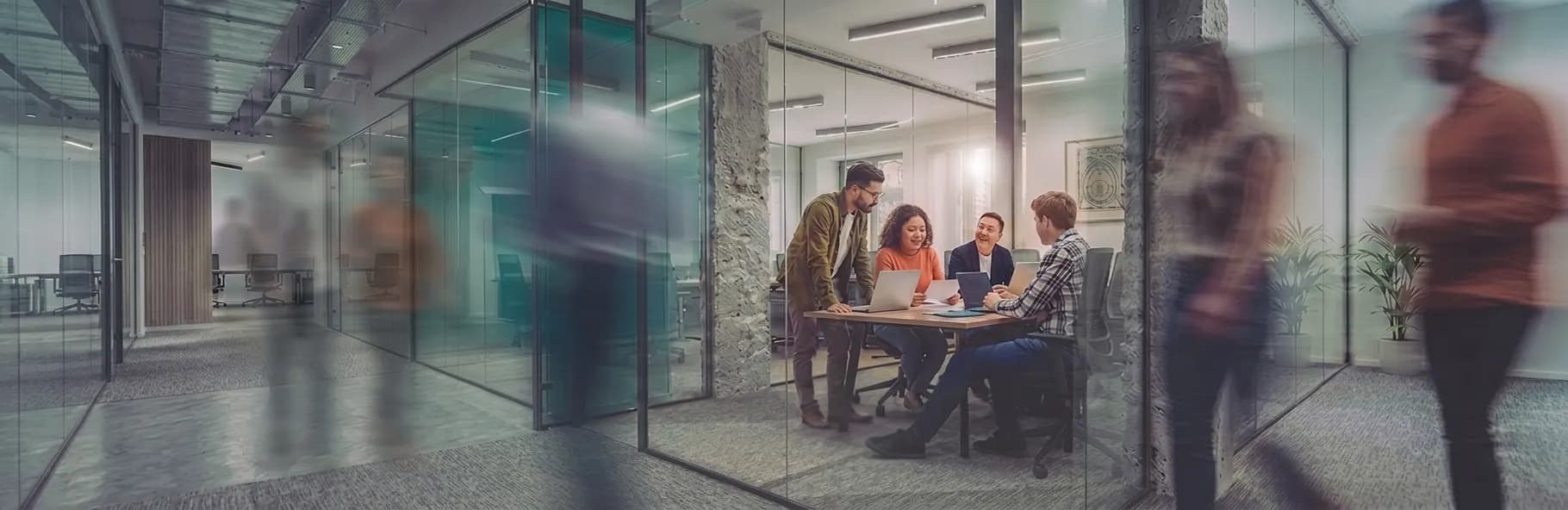 A team meeting in a modern glass-walled office — colleagues seated at a table reviewing laptops together while others walk past in motion-blurred foreground.